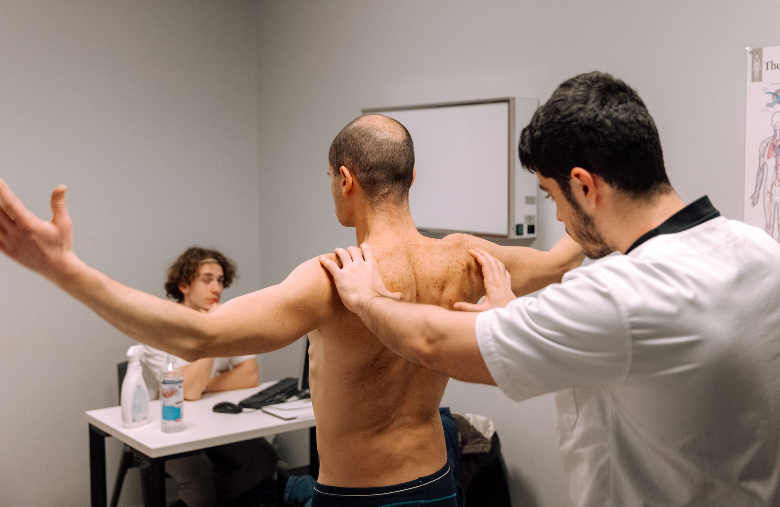 A healthcare professional in a white coat examining the back and shoulders of a shirtless man with arms extended in a clinical setting while another person sits at a desk in the background.