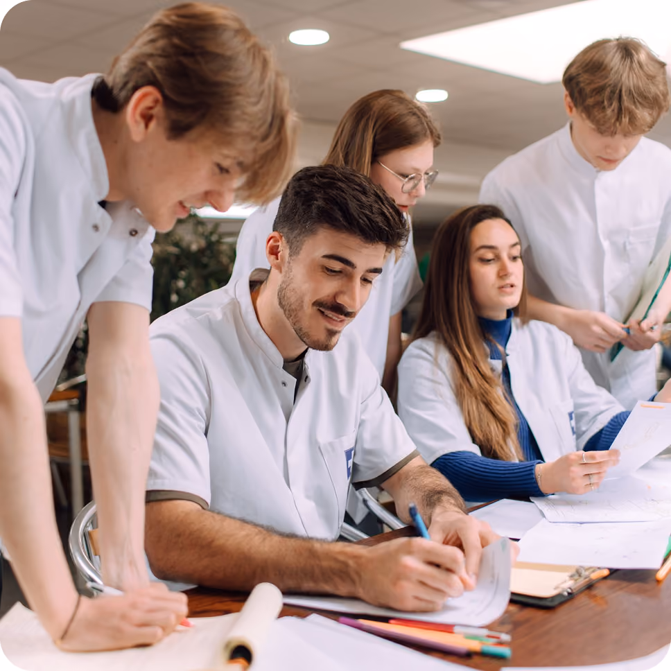 Five young adults in white coats engaged in collaborative work at a table with papers and colored pencils, focused on writing and reviewing documents indoors under ceiling lights.