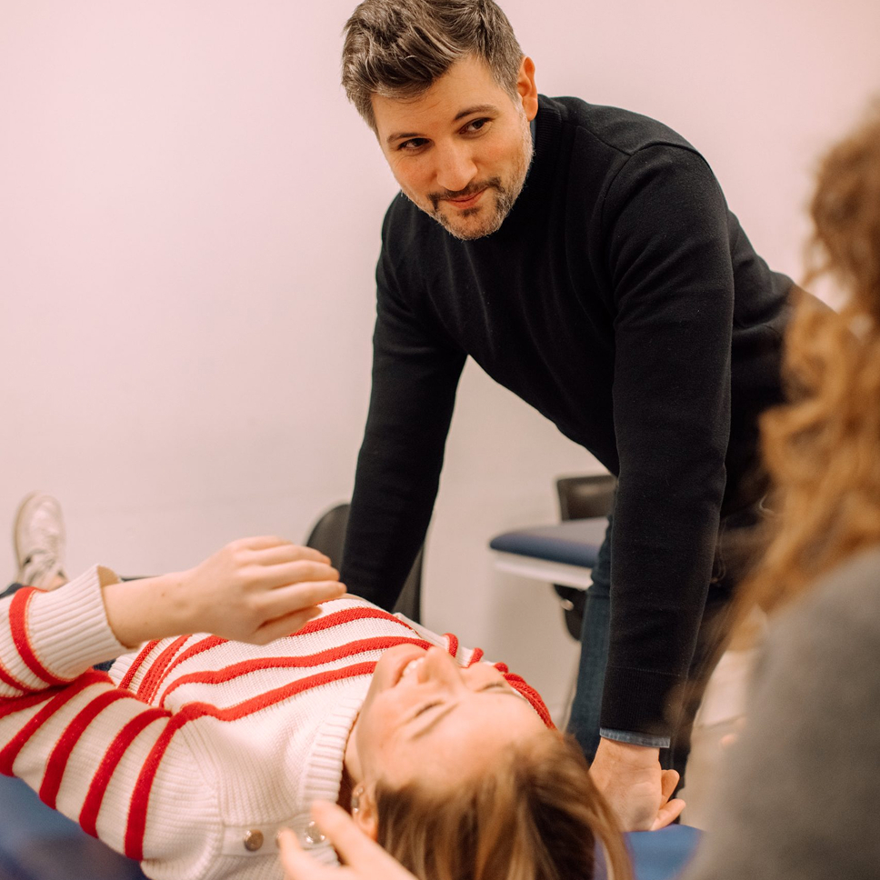 A man in a black sweater leans forward looking attentively at a woman lying down wearing a white sweater with red stripes while another person with curly hair looks on.