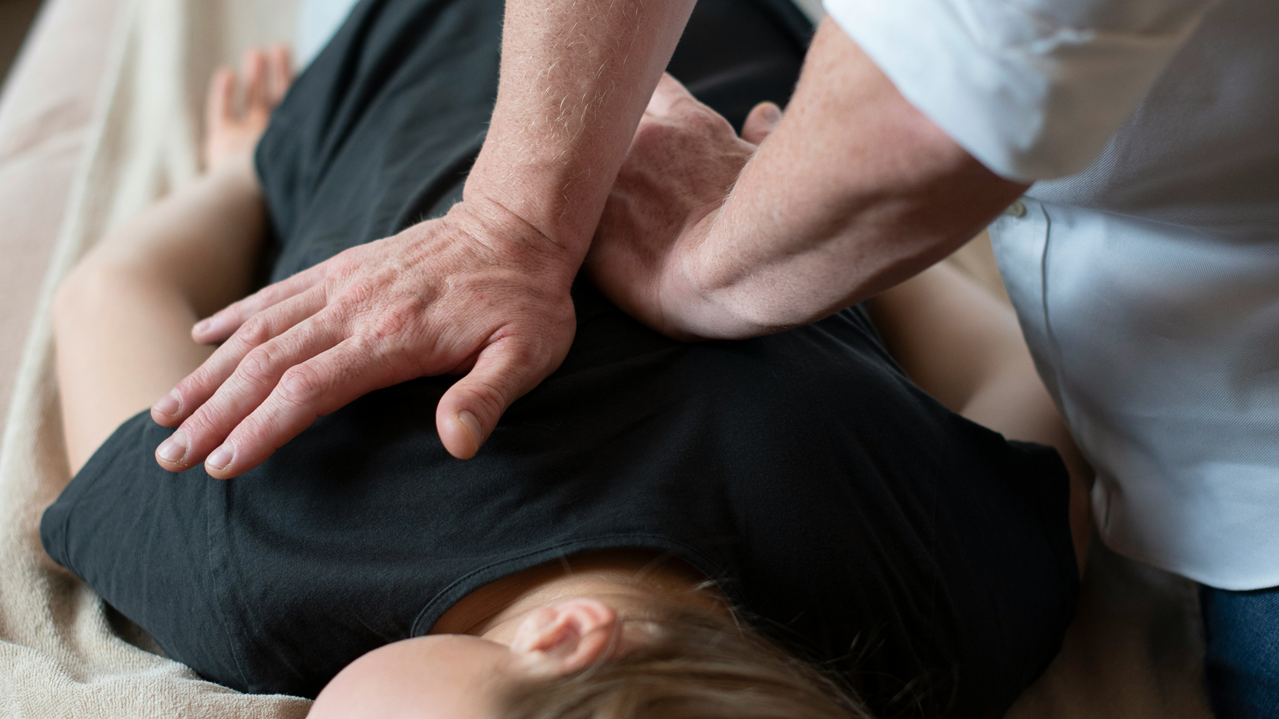 A person lying face down on a bed covered with a beige towel receiving a chiropractic adjustment on their back from another person using both hands.