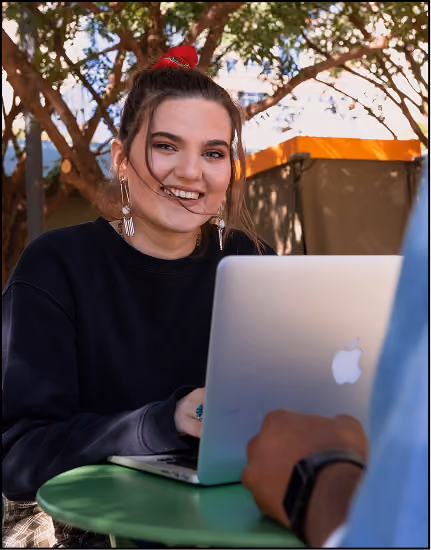 Smiling young woman with a red scrunchie and earrings sitting outdoors at a green table, using a silver Apple laptop, with another person partially visible in front of her.