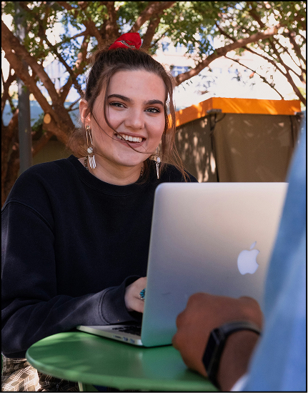 Smiling young woman with a red scrunchie and earrings sitting outdoors at a green table, using a silver Apple laptop, with another person partially visible in front of her.