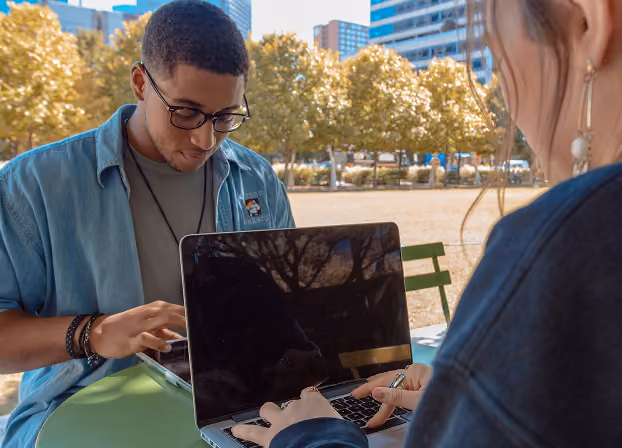Two young adults sitting at a green table outdoors, one man wearing glasses looks down at a laptop and phone, a woman’s hands type on the laptop keyboard, trees and buildings in background.
