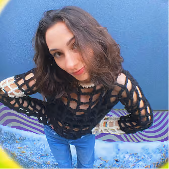 Woman with shoulder-length wavy brown hair wearing a black open-knit sweater and blue jeans, standing in front of a blue wall with a textured ground.
