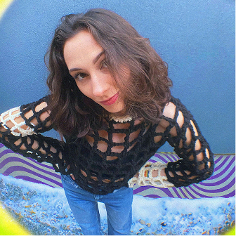 Woman with shoulder-length wavy brown hair wearing a black open-knit sweater and blue jeans, standing in front of a blue wall with a textured ground.