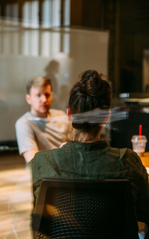 Two people in conversation across a table in a modern office space; a woman with her hair in a bun seen from behind and a man sitting in front of her, slightly blurred.