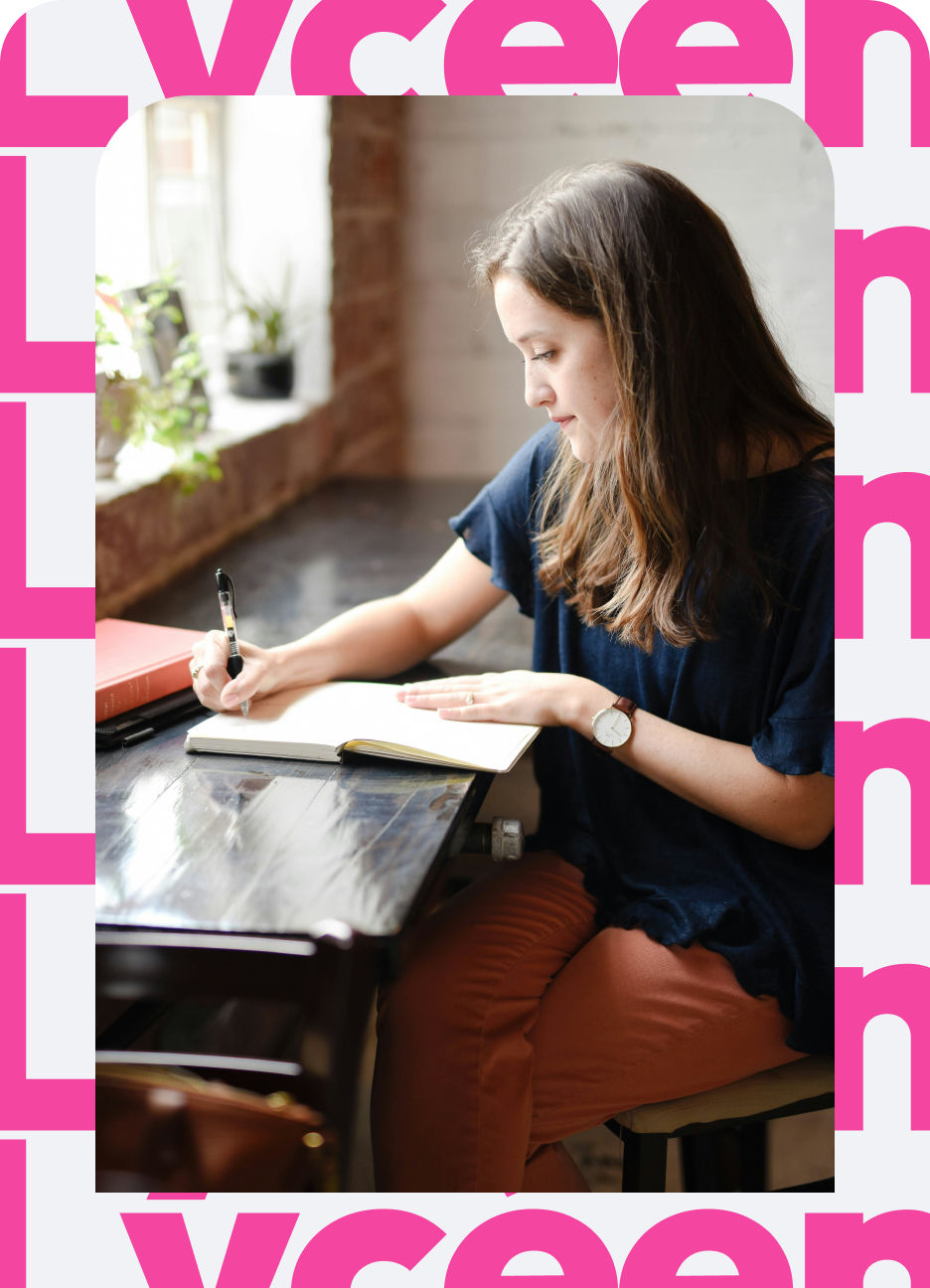 Young woman with long brown hair sitting at a dark wooden table by a window, writing in an open notebook with a black pen, wearing a navy blue top, orange pants, and a wristwatch.