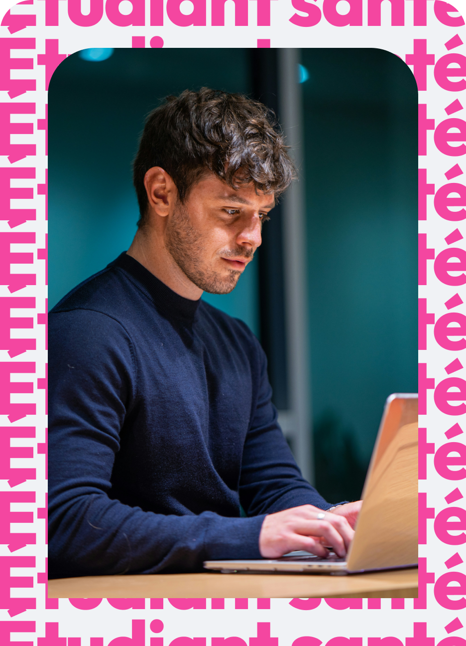 Young man with short dark hair and a beard wearing a navy blue sweater working on a laptop at a wooden table indoors with a dark teal background.