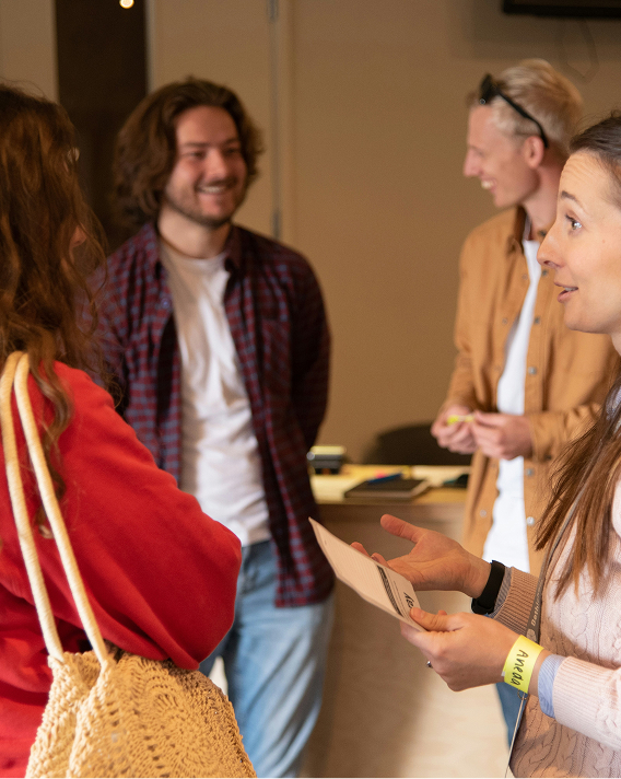 Four young adults engaged in conversation indoors; a woman in a pink sweater holds a paper and gestures while a woman in a red jacket listens with a woven bag, with two men smiling in the background.