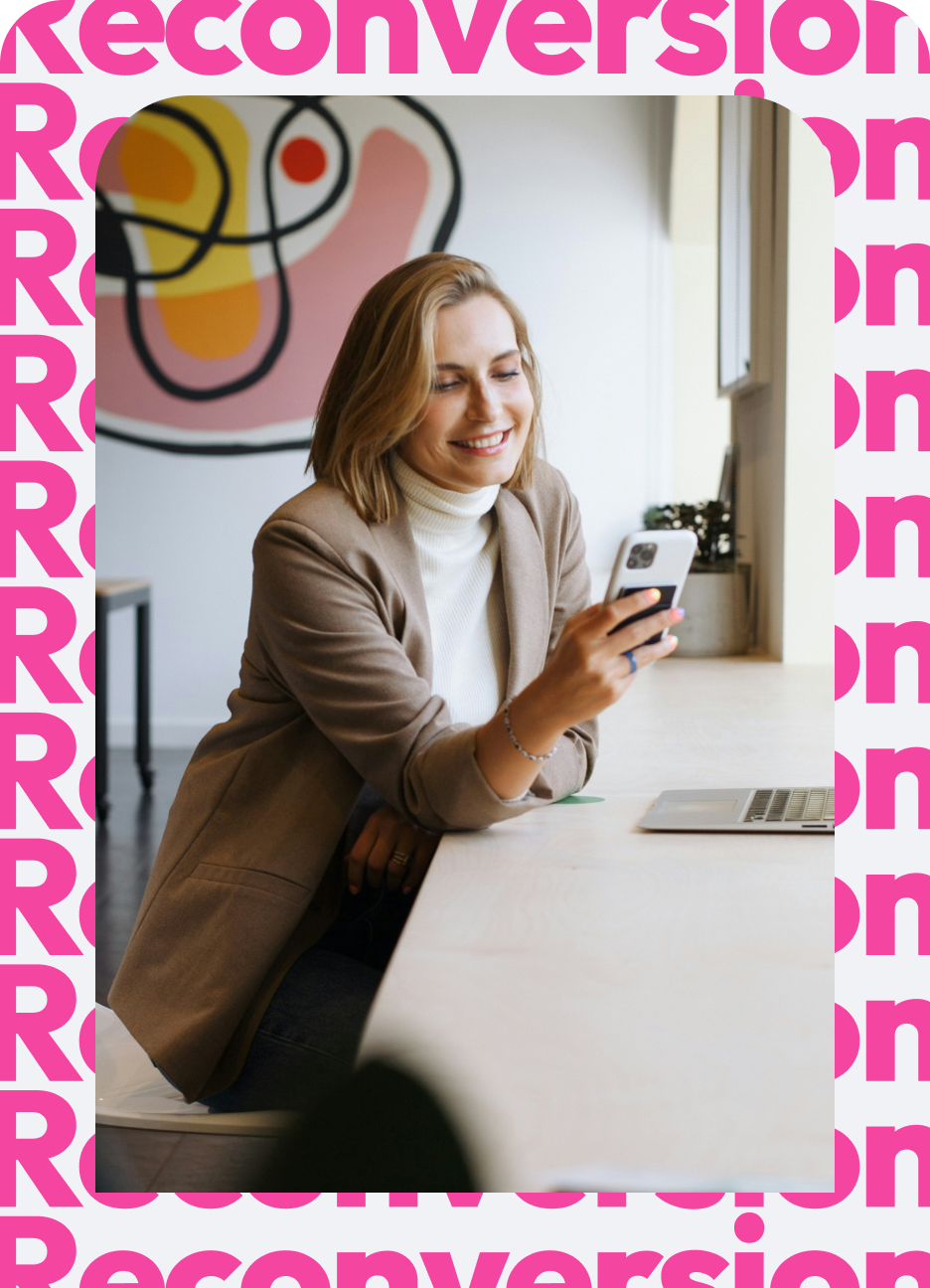Smiling woman wearing a beige blazer and white turtleneck sitting at a light wooden desk by a window, looking at her smartphone with a laptop in front of her.