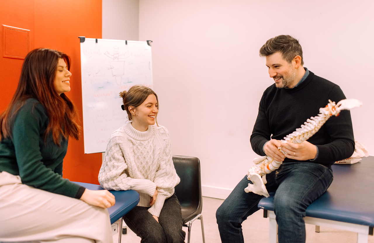 A man holding a spine model is seated on an examination table, explaining to two women sitting nearby, one in a green sweater and the other in a white cable-knit sweater, in a clinical room with a red wall and a flip chart in the background.