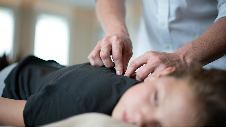 A person receiving a back massage or chiropractic adjustment while lying face down, with the practitioner's hands pressing on the upper back.