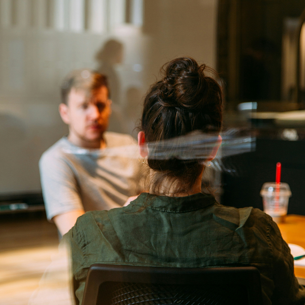 Two people sitting across from each other at a table in a casual indoor setting, with one person visible from behind and the other slightly blurred in the background.