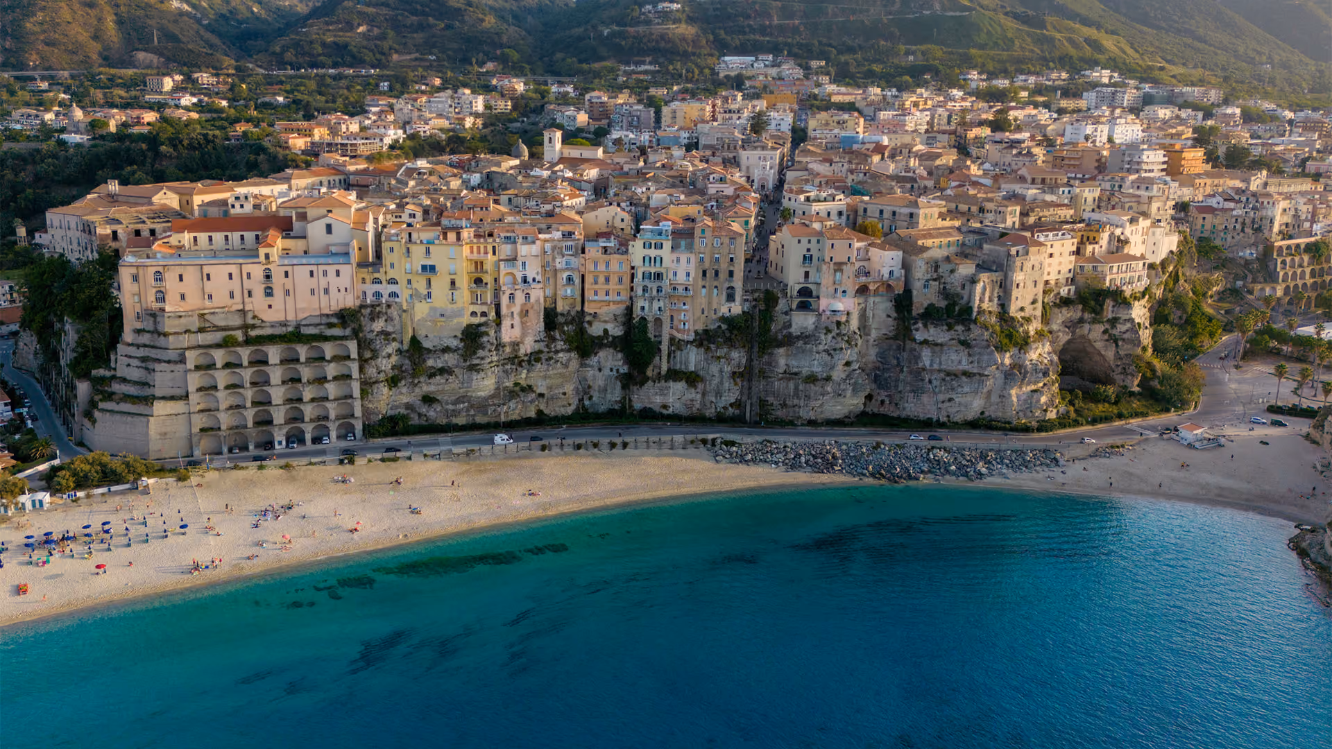 Cliffside town with pastel buildings overlooking a sandy beach and turquoise sea under mountains.