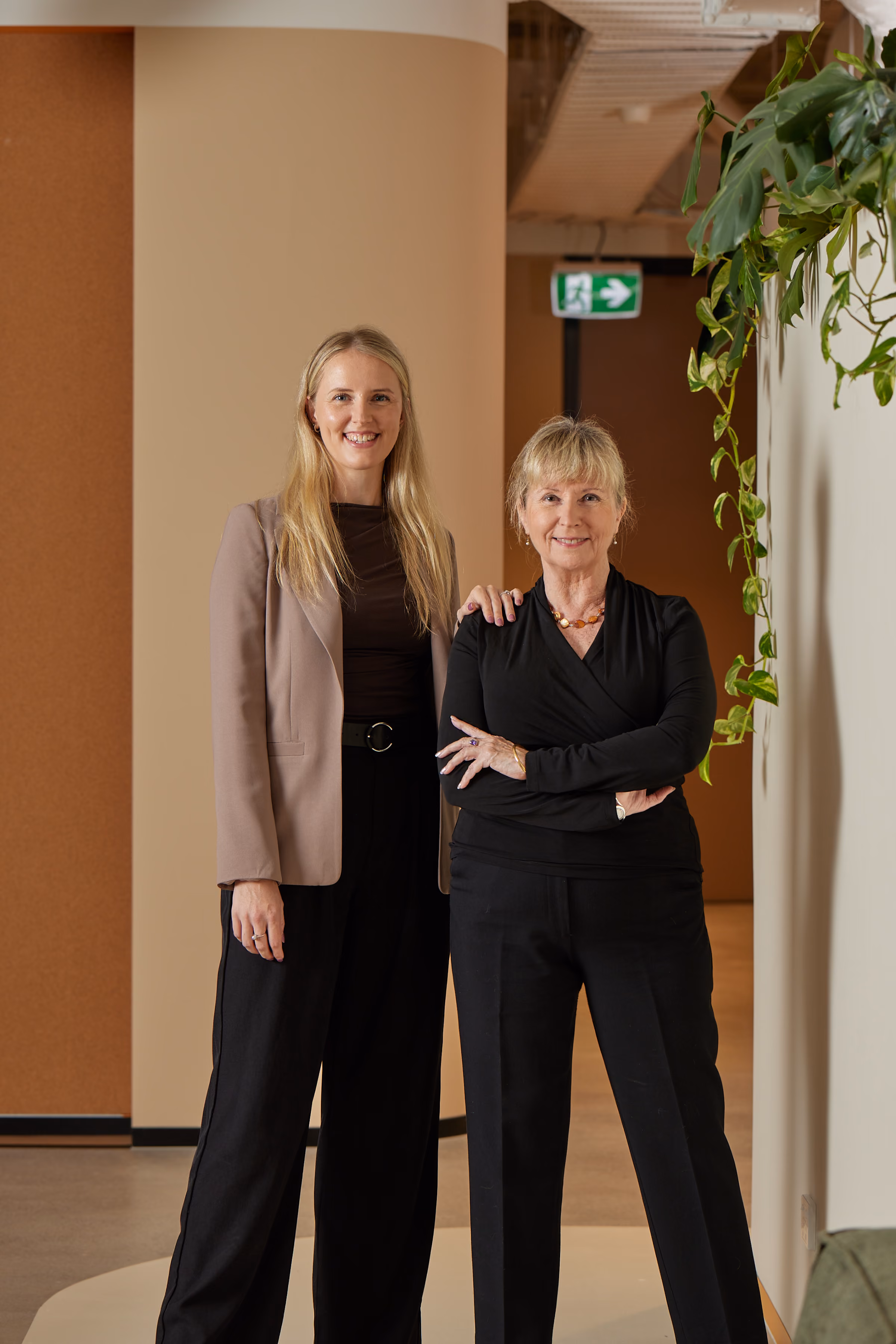 Two women standing together indoors, one with blonde hair in a blazer, the other in black with arms crossed.