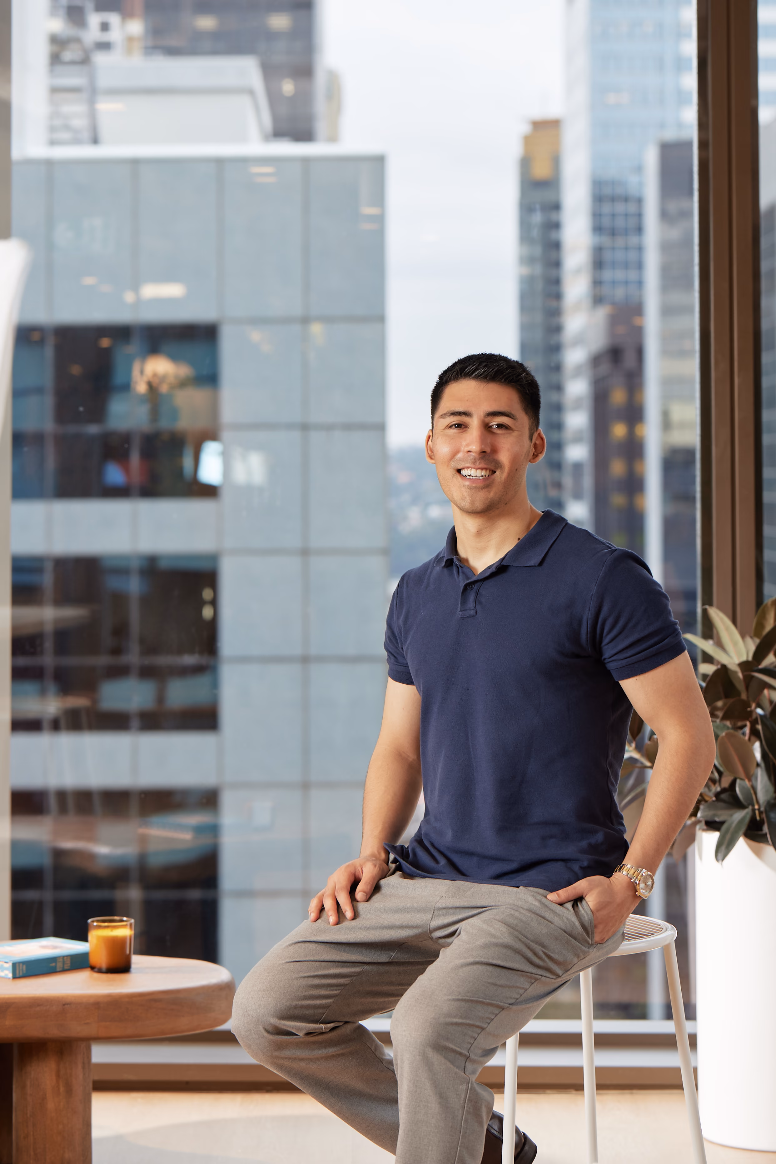 Smiling man in a navy polo shirt and gray pants sitting on a stool by a window with skyscrapers outside.