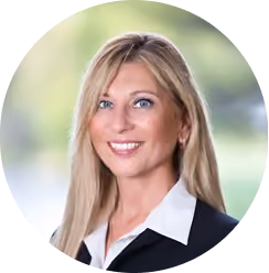 Portrait of a smiling blonde woman with blue eyes wearing a black blazer and white shirt.