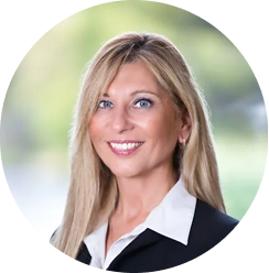 Portrait of a smiling blonde woman with blue eyes wearing a black blazer and white shirt.