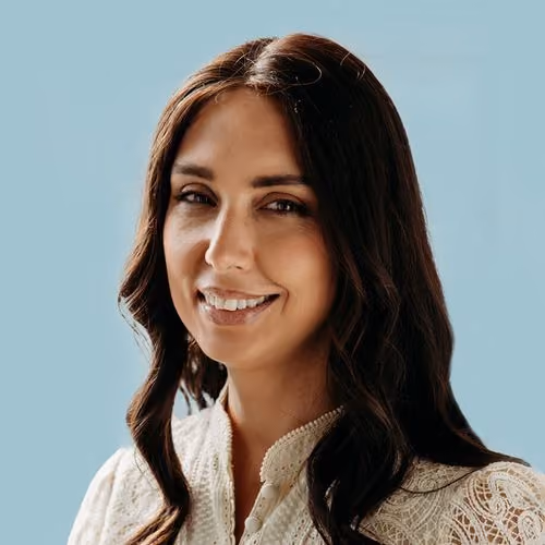 Smiling woman with long dark hair wearing a white lace blouse against a light blue background.
