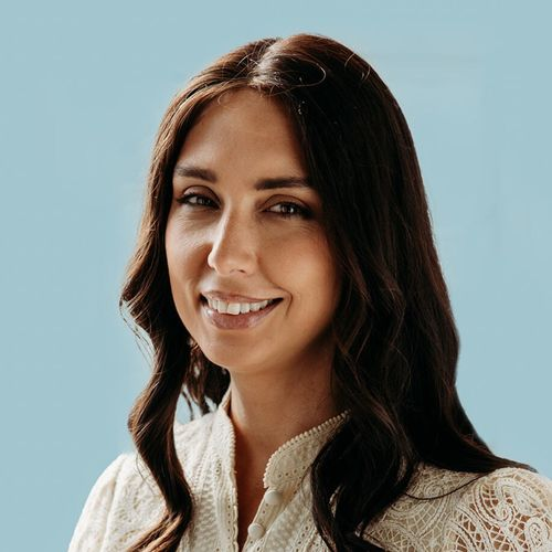 Smiling woman with long dark hair wearing a white lace blouse against a light blue background.