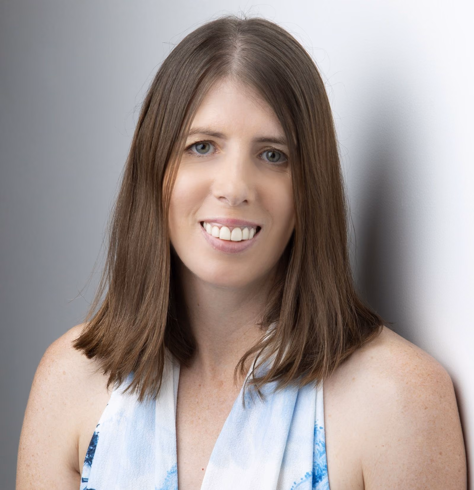 Smiling woman with shoulder-length brown hair wearing a light blue patterned sleeveless top.