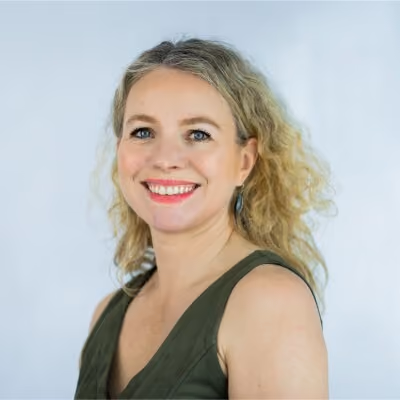 Smiling woman with curly blonde hair wearing a green sleeveless top and earrings against a light background.