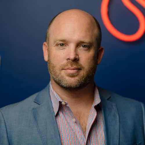 Portrait of a bald man with beard wearing gray blazer and checkered shirt against a blue background.