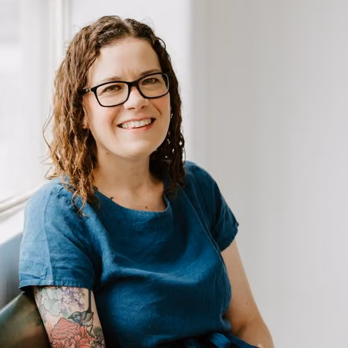Smiling woman with glasses, curly hair, blue shirt, and floral tattoo on her arm sitting by a window.
