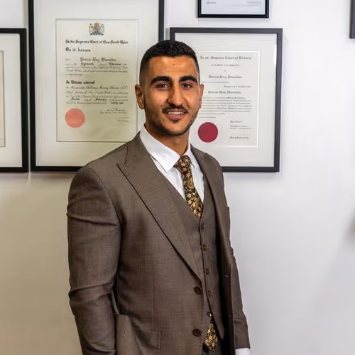 Man in a brown suit and floral tie standing in front of framed certificates on a wall.