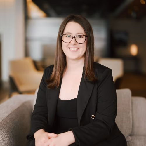 Smiling woman with glasses and brown hair wearing a black blazer seated on a gray sofa.