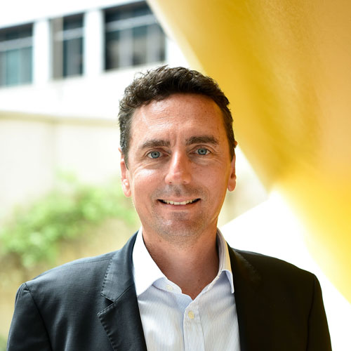 Man with short dark hair wearing a black blazer and white shirt, smiling outdoors by a yellow structure.