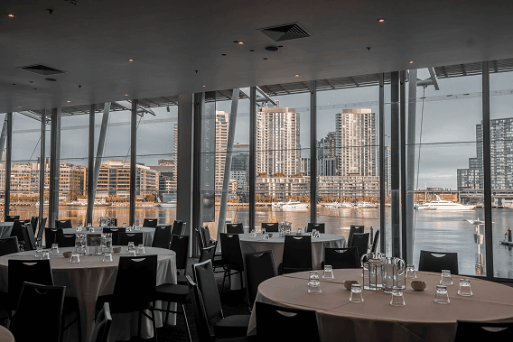 Empty restaurant dining area with round tables set with glasses overlooking waterfront city skyline.