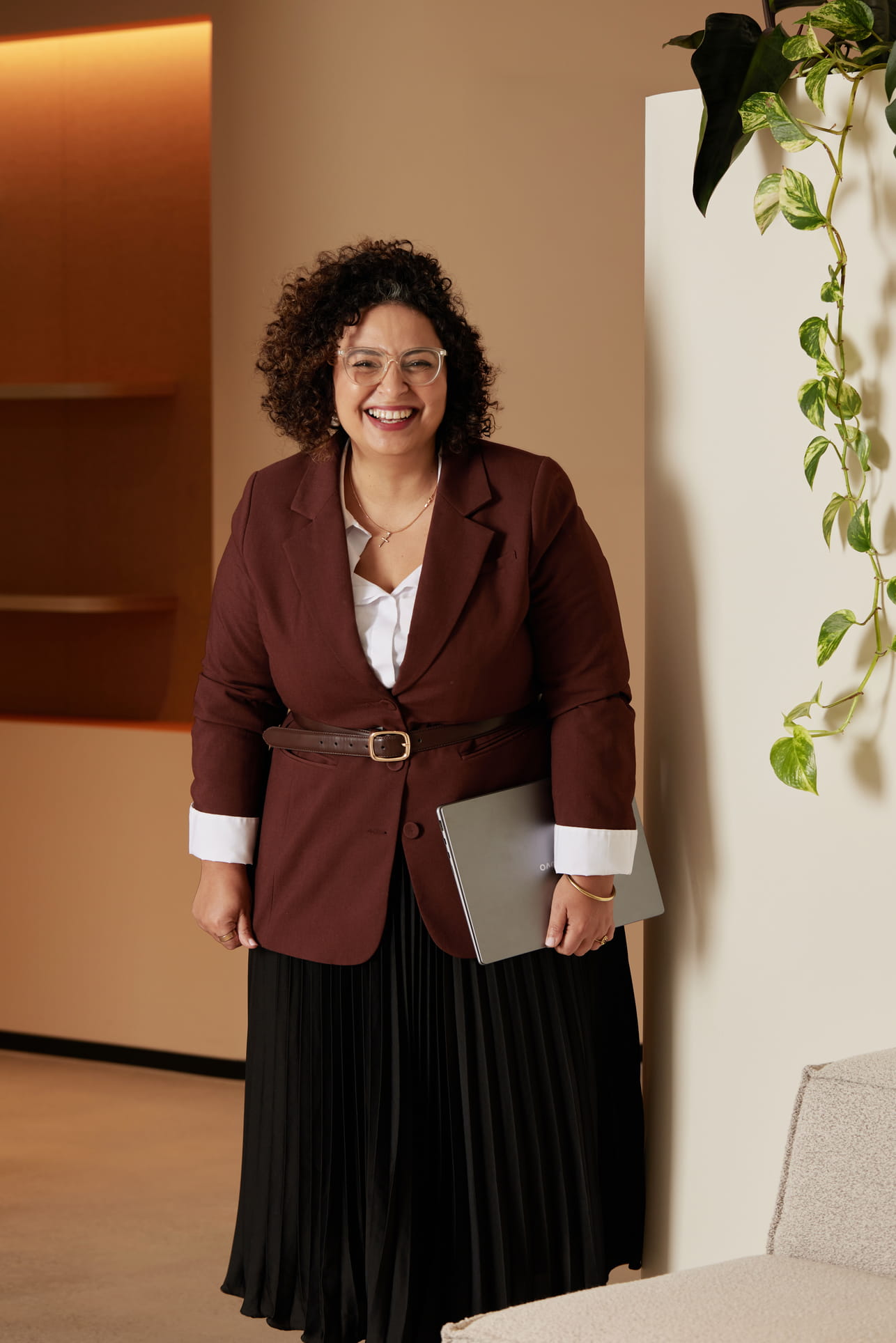 Smiling woman with curly hair wearing glasses, brown blazer, and black skirt, holding a laptop.