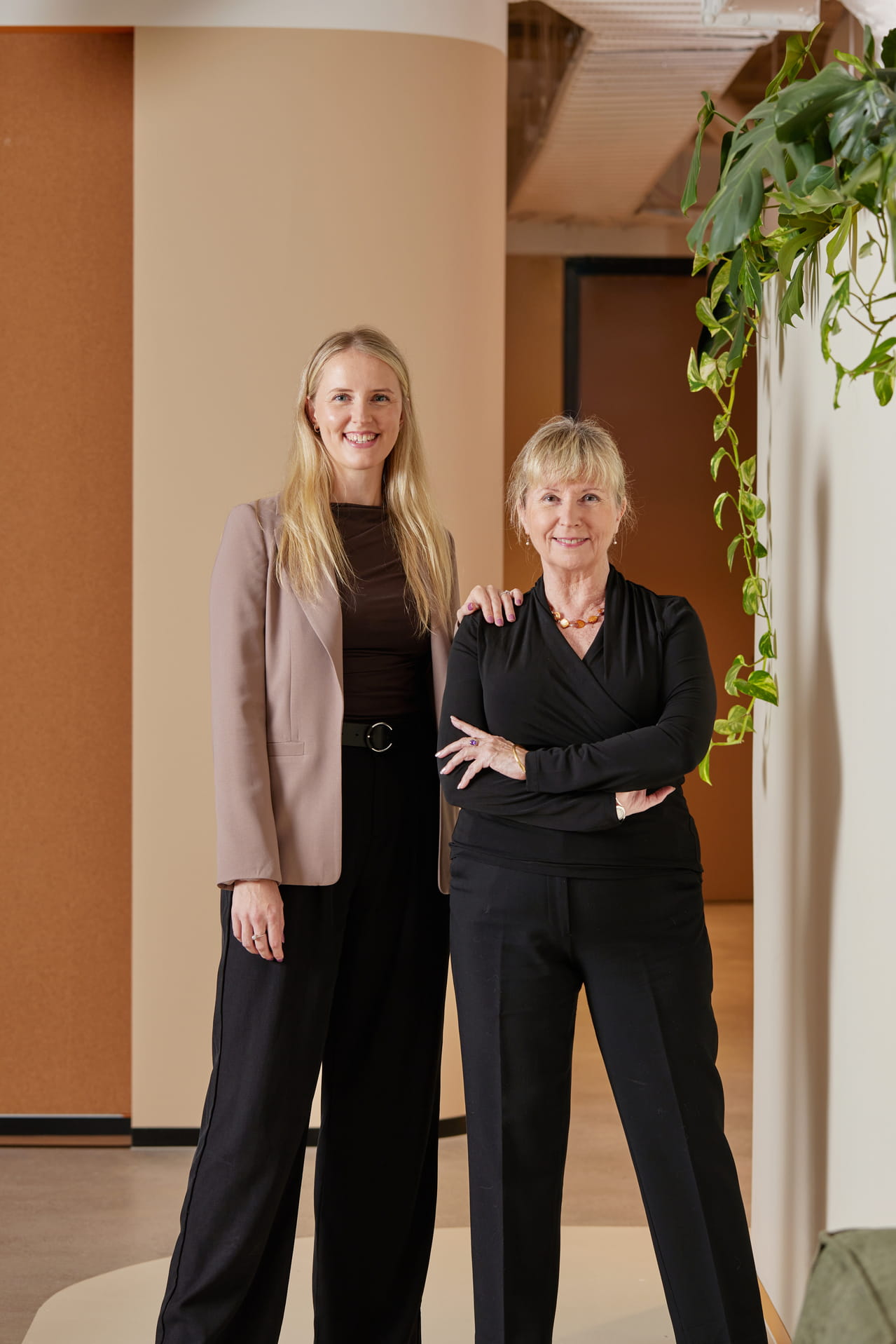 Two women smiling, standing indoors next to each other with one woman’s hand on the other's shoulder.