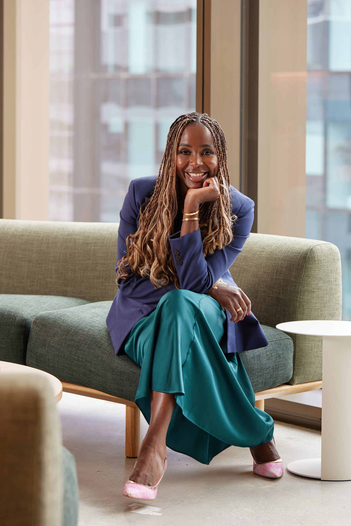 Smiling woman with braids wearing a blue blazer and teal skirt sitting cross-legged on a couch.