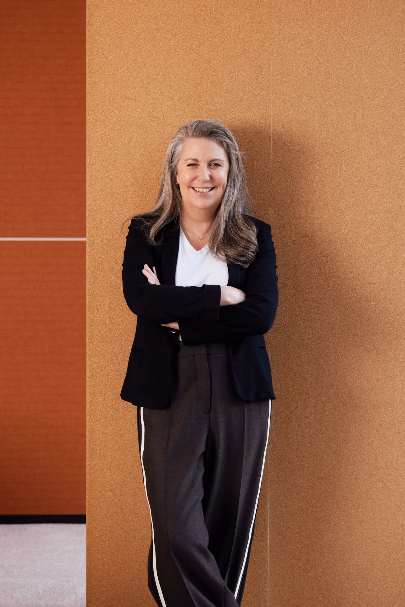 Smiling woman with gray hair in black blazer and striped pants leaning against a tan wall.