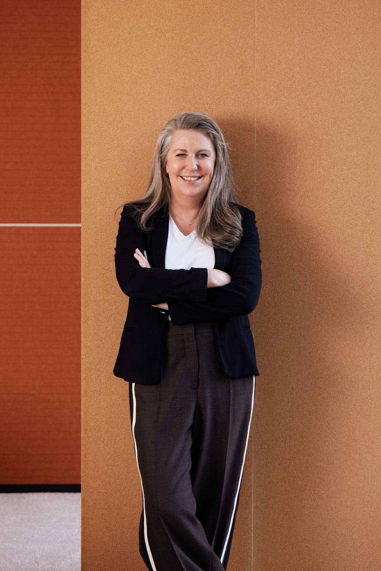 Smiling woman with gray hair in black blazer and striped pants leaning against a tan wall.