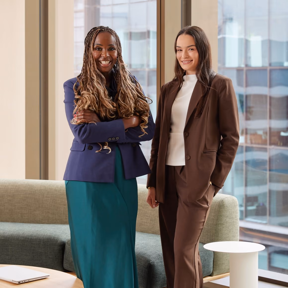 Two professional women smiling and standing in an office lounge area with large windows.