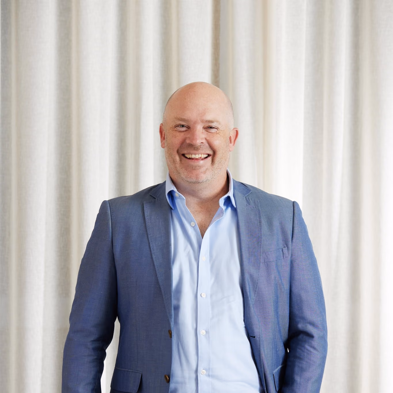 Middle-aged bald man smiling, wearing a blue blazer and light blue shirt, standing before cream curtains.