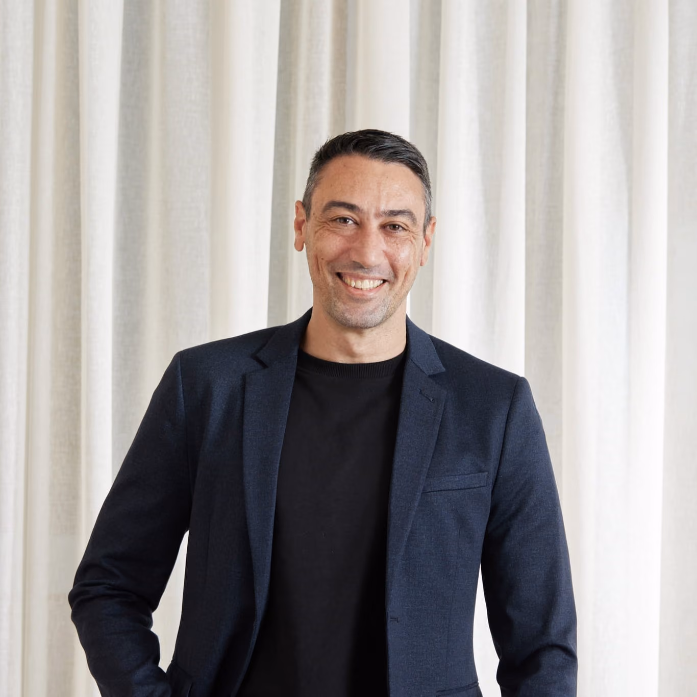 Smiling man with short dark hair wearing a dark blazer and black shirt in front of light curtains.
