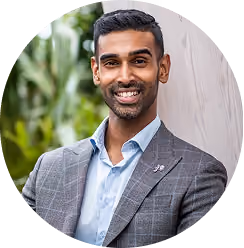 Smiling man in a plaid blazer and light blue shirt standing outdoors near wood and greenery.