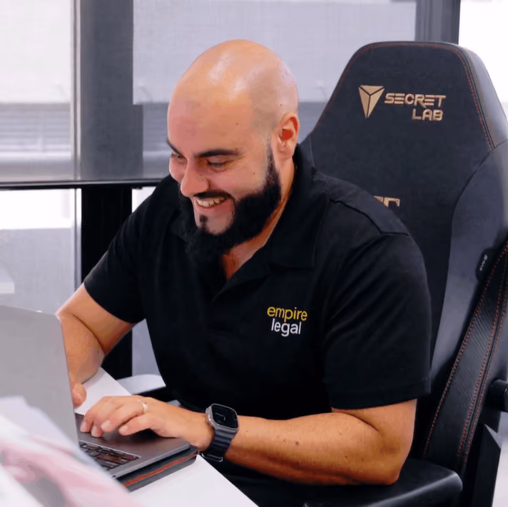 Smiling bald man with a beard working on a laptop, wearing a black Empire Legal polo shirt.