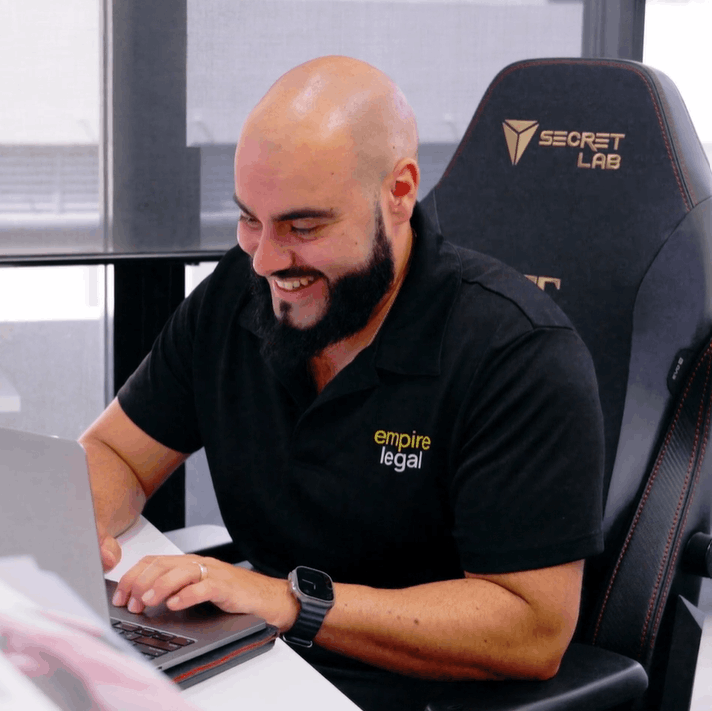 Smiling bald man with a beard working on a laptop, wearing a black Empire Legal polo shirt.