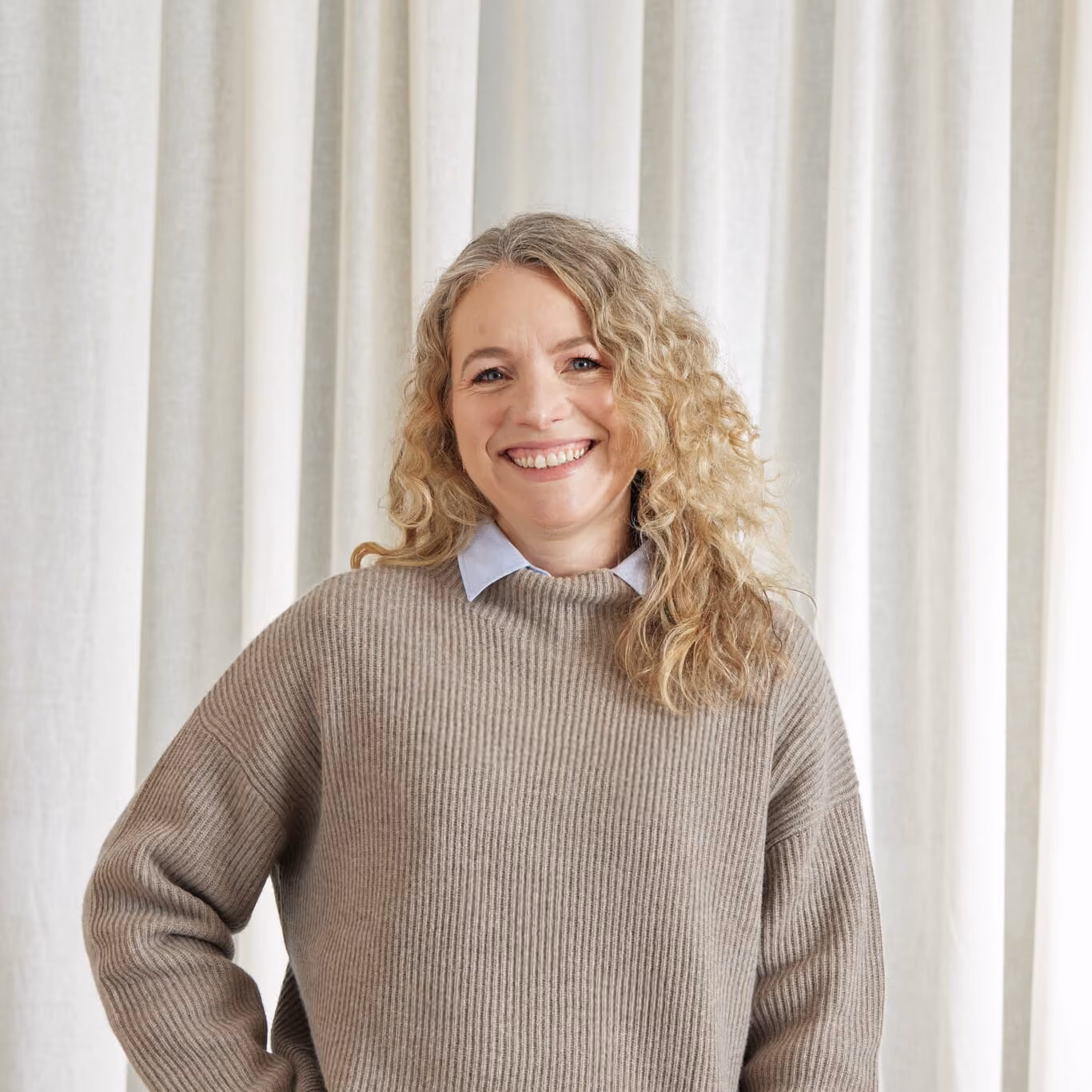 Smiling woman with curly blonde hair wearing beige sweater and blue collared shirt in front of curtains.