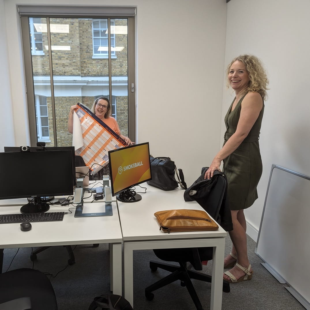 Two women in an office: one holds a large calendar, the other stands smiling by a desk with monitors.
