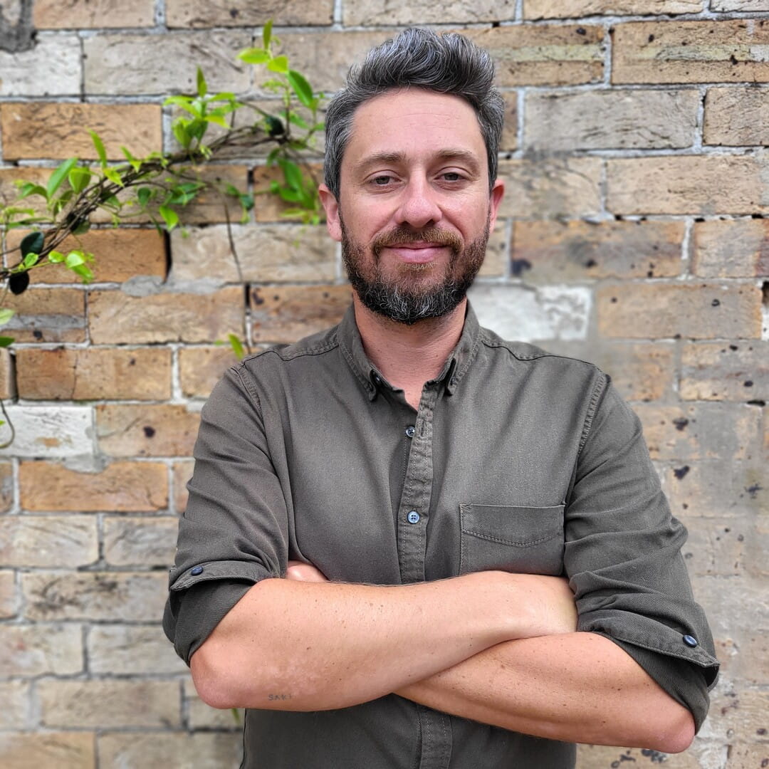 Man with dark gray hair and beard, wearing a rolled-up sleeve button shirt, standing with arms crossed.