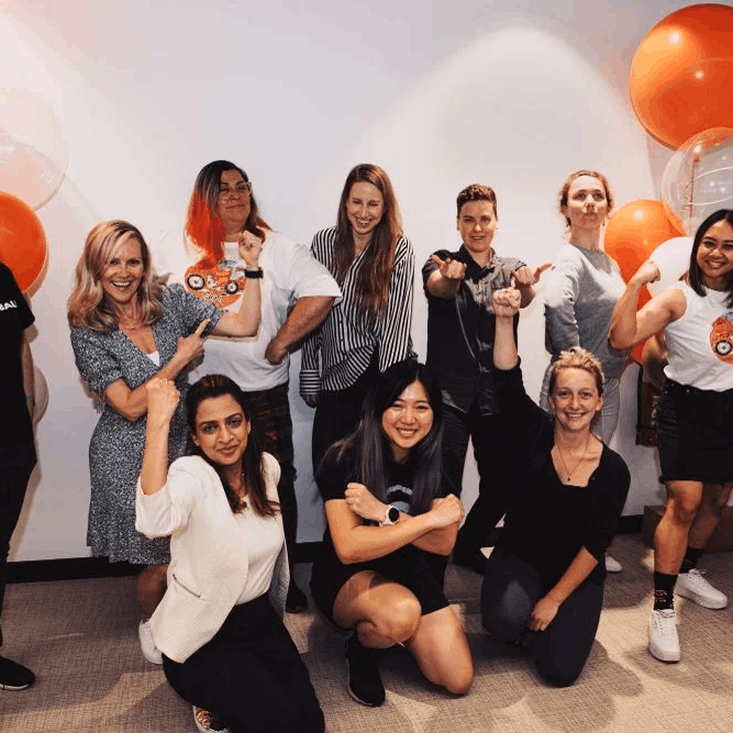 Group of diverse smiling people posing with flexed arms, orange and white balloons in background.
