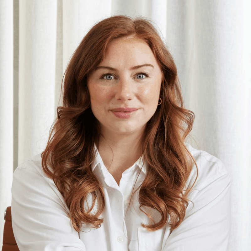 Smiling woman with long red hair wearing a white shirt sitting in front of white curtains.