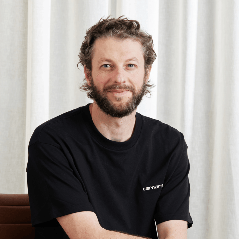 Man with beard and curly hair wearing a black Carhartt t-shirt, sitting and smiling at the camera.