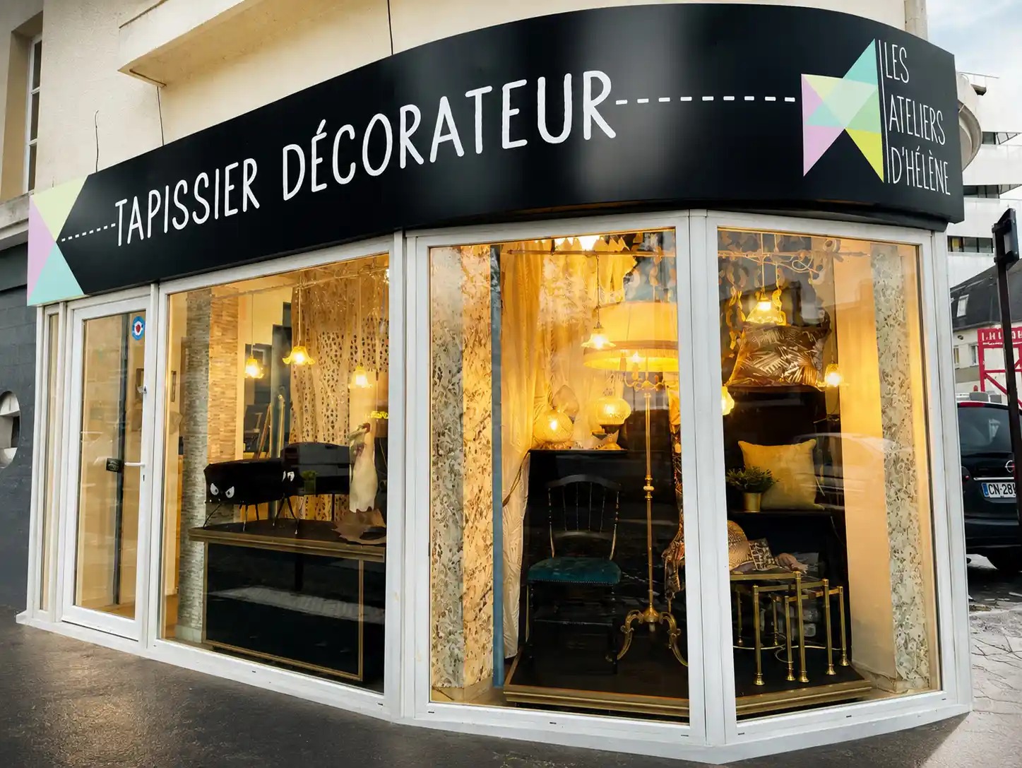 Storefront of Les Ateliers d'Hélène, a decor and upholstery shop with lit lamps and decorative items in windows.